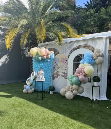 Outdoor birthday party decoration with a balloon arch, tiered cake, and palm tree on a green lawn.