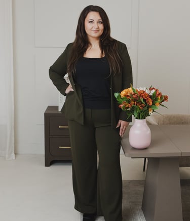 Adriana in dark green blazer and trousers standing by a desk with autumn flowers.