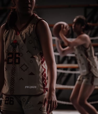 A female basketball player in a custom patterned jersey stands on court while a teammate shoots a ball.