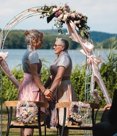 Zwei Bräute halten sich bei der freien Trauung unter einem Blumenbogen an den Händen. Foto: Joel Pingel.