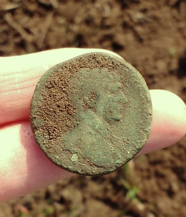 A large Roman coin with green patina, held out on two fingers of the finder.