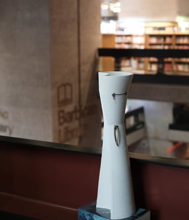 A tall elegant white porcelain vessel, with bronze knot details, on a shelf in the Barbican Library