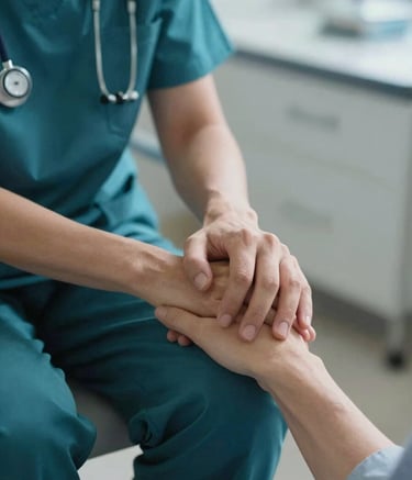 A close-up photography shot of a healthcare provider in dark teal medical attire holding a patient's hand comfortingly. The scene is set in a warm, North American clinical environment with soft, empathetic lighting and light blue-grey tones.