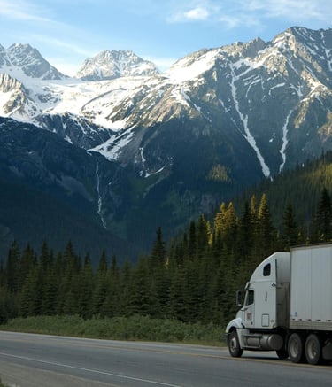 A semi-truck driving on a scenic highway through snow-capped mountains and evergreen forests.