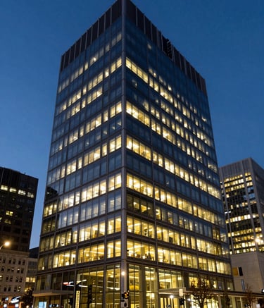 A sharp, wide-angle photograph of a contemporary office building in a North American city at dusk, glowing with warm internal lights against a dark blue sky, representing modern and clean geometric architecture.