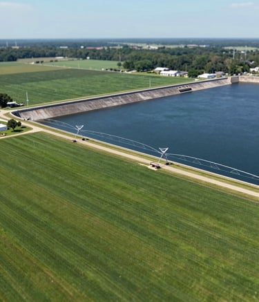 A wide-angle aerial shot of a well-organized farm in MS, showing a modern dam and irrigation infrastructure. The composition is clean and expansive, reflecting a consolidated engineering firm. Colors focus on the lush green of the plantation (#2E5F49) and the sophisticated dark blue of the water (#0F2027).