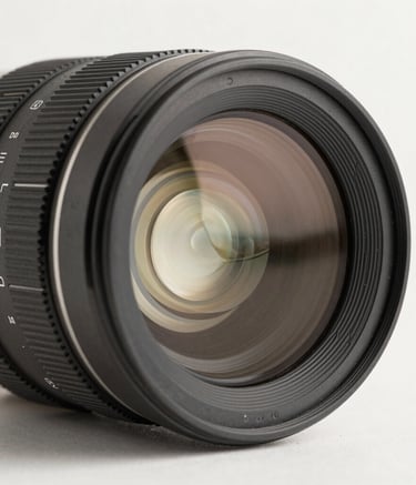 A macro, high-detail photograph of a high-end cinema lens showing intricate glass elements and focus markings. The lighting is clean and professional with neutral tones, set against an off-white background in a South American / Brazilian production studio.