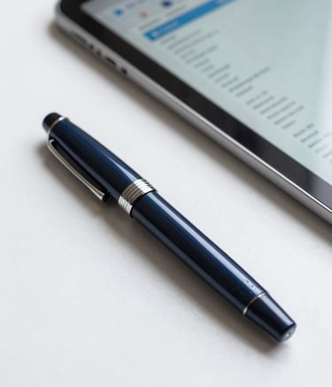 Close-up of a high-end fountain pen resting on a clean desk next to a blurred tablet displaying analytical interfaces. The color palette includes dark slate blue and soft sky blue highlights in a clean, professional setting.