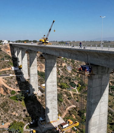 A wide aerial shot of a massive concrete bridge under construction across a deep valley in a Mexican landscape. The scene features heavy machinery, steel blue sky, and technicians wearing navy blue and orange safety gear. Professional photography, high contrast, corporate style.