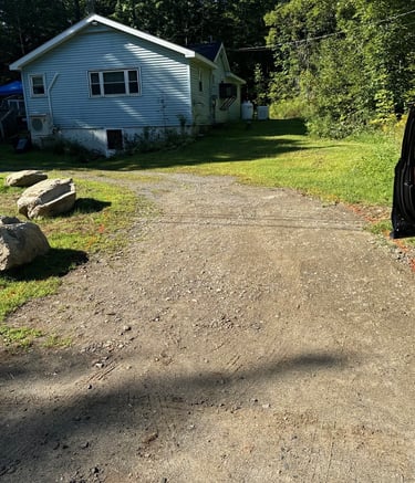 A gravel driveway leading to a light blue house surrounded by green trees and large decorative landscape rocks.