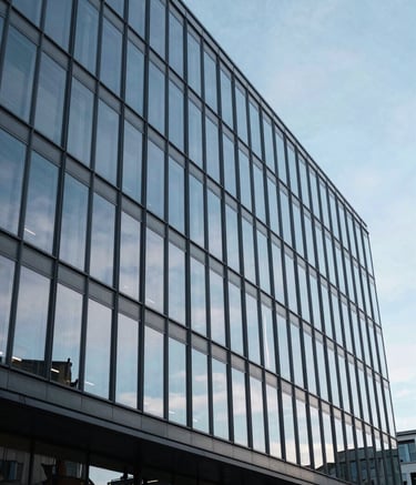 A wide-angle photography shot of a modern, clean glass building facade in Cardiff, Wales, reflecting a soft blue sky. The composition is professional and authoritative, using a palette of slate blue and light grey to suggest stability.
