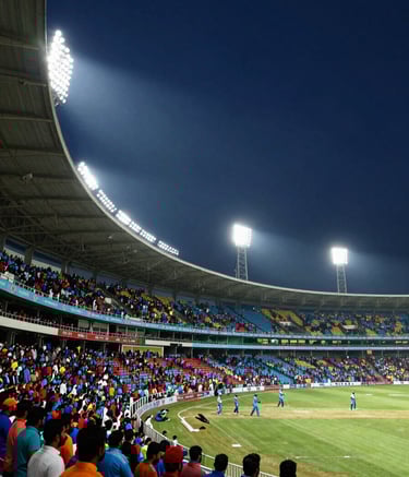 A wide-angle photography shot of a high-energy cricket stadium in India during a night match. The stands are filled with South Asian / Indian fans in colorful attire, illuminated by bright white floodlights. The sky is a deep navy blue, creating a high-contrast and exciting atmosphere.
