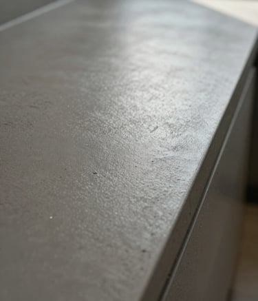 A close-up photograph of a professional decorative concrete finish. The texture shows a high-end polished surface in muted steel grey, reflecting soft natural light. The composition is a sharp angle shot highlighting the smooth, industrial craftsmanship.