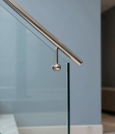 Close-up architectural photography of a minimalist glass handrail with polished stainless steel fixtures. Set in a luxury Riyadh villa interior with Middle Eastern / Gulf aesthetics. High-end precision finishing, sharp focus, professional studio lighting, with a palette of pale silver-blue and slate blue.