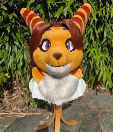 Custom orange feline fursuit head with striped ears and brown hair displayed outdoors.
