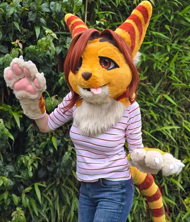 Person in a yellow and orange striped feline fursuit waving near green foliage.