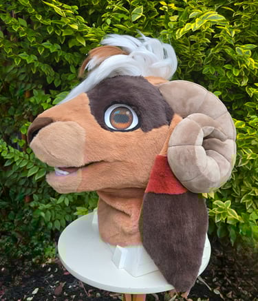 Custom brown ram fursuit head with curved horns and white hair displayed outdoors against green leaves.