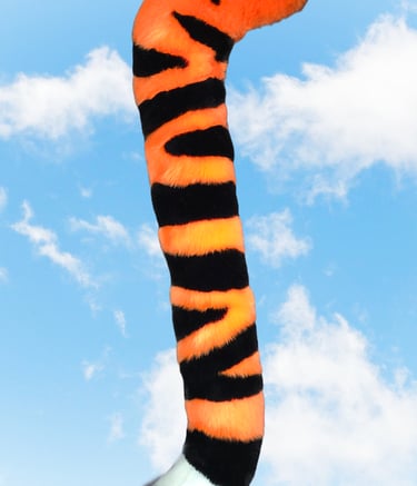 Handmade orange and black tiger faux fur tail for furry cosplay accessories with a white tip against a blue sky.