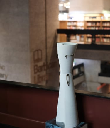 A tall elegant white porcelain vessel, with bronze knot details, on a shelf in the Barbican Library