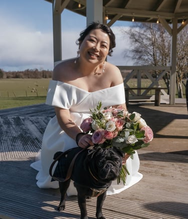 Smiling bride in a white wedding dress holding a rose bouquet next to her black pug dog.