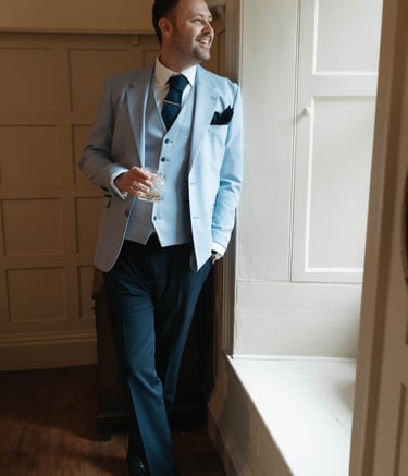 Groom in light blue three-piece windowpane suit holding whiskey by a window in glasgow.