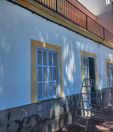 White stucco Mediterranean house with blue shutters and a metal ladder on the sidewalk.
