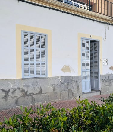 Traditional white Mediterranean house facade featuring light blue wooden shutters and a sidewalk with green bushes.