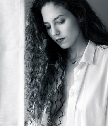 Portrait fine art en noir et blanc d’une femme aux cheveux longs, posant près d’une fenêtre