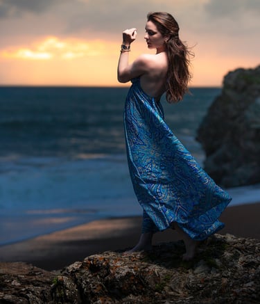 Portrait créatif d’une femme en robe bleue posant sur des rochers face à la mer au coucher du soleil