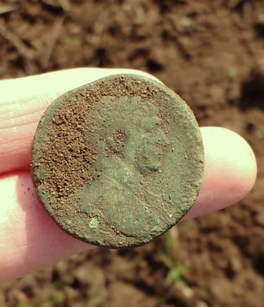 A large Roman coin with green patina, held out on two fingers of the finder.