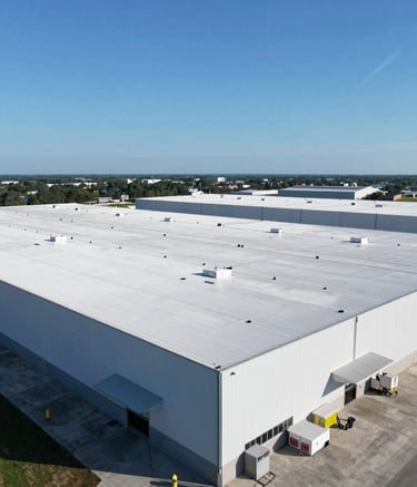 A wide-angle aerial drone photograph of a massive industrial warehouse complex with a pristine white TPO flat roof in North American / US. The composition shows the scale and cleanliness of the roofing work against a clear blue sky, emphasizing reliability and professional capacity.