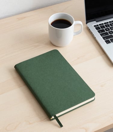 A minimalist Scandinavian-inspired desk setup with a clean crisp parchment wooden surface. A matte forest green notebook, a white ceramic mug of coffee, and a slim modern laptop are arranged neatly. The lighting is soft and natural, suggesting a professional yet calm morning workflow in a bright studio.