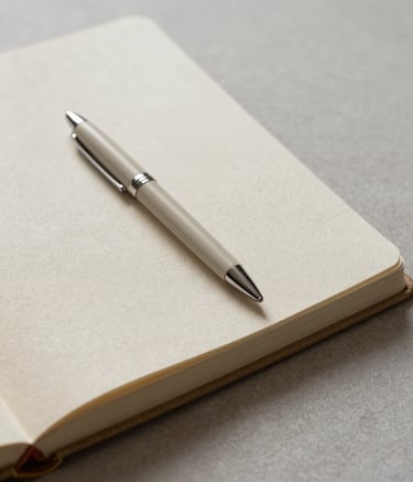A refined, close-up photograph of an open linen-covered notebook and a premium minimalist pen resting on a muted light gray textured surface. The scene is set in a professional North American / European studio with soft, diffused natural light, emphasizing a calm and intellectual mood in warm beige tones.