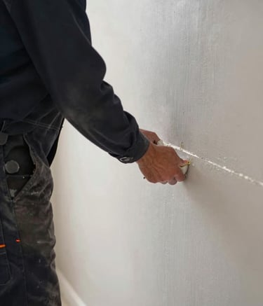 A close-up photograph of a professional drywaller in a modern North American / US home, carefully applying joint compound to a seam. The worker is wearing a clean Dark Navy uniform. The lighting is bright and even, highlighting the smooth texture of the Pearl Grey drywall. The composition is a focused side-angle shot.