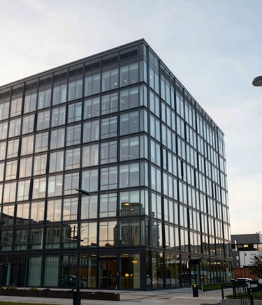A wide-angle photography shot of a modern, established office building in a United Kingdom industrial park during a bright morning. The architecture is clean and professional, with reflection on glass windows. The sky is a soft off-white and light blue.