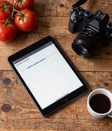A top-down lifestyle photograph of a rustic wooden desk in a North American / Western European office. On the desk are a tablet showing a content calendar, a professional camera, fresh vine-ripened tomatoes, and a cup of espresso. The mood is professional and organized with a cozy, artisanal touch.