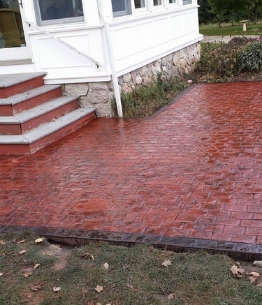 Custom red stamped concrete patio with decorative stone borders installed next to a white house.