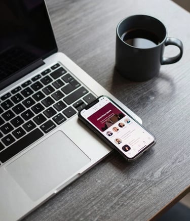 A top-down professional shot of a graphite grey wooden desk. On the desk sits a sleek laptop, a smartphone displaying a social media dashboard with deep red accents, and a charcoal black coffee mug. The lighting is modern and bright.