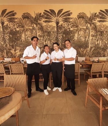 Restaurant service team posing in a tropical-themed dining room with palm tree wallpaper.