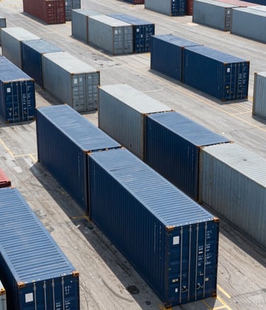 A high-angle professional photograph of a bustling shipping port in the Middle Eastern / Gulf region. Neatly organized rows of cargo containers in navy and gray reflect efficiency and global scale. The bright midday sun creates sharp, clean shadows, emphasizing a modern logistics operation.