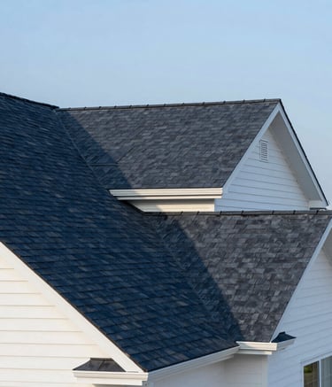 A wide-angle, professional photograph of a beautifully shingled residential roof in a North American / US suburban neighborhood. The shingles are a crisp Dark Navy, contrasting with the Off-White exterior of the house. The sky is a clear Soft Blue, suggesting a reliable and sturdy structure under bright daylight.