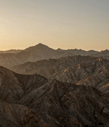 A panoramic photograph of a rugged mountain range in the North American wilderness during the golden hour. The landscape is bathed in muted tan and gold-brown light, emphasizing texture and depth without any human presence. Professional and minimalist aesthetic.