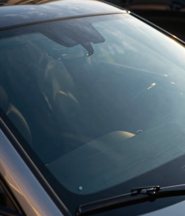 A close-up high-resolution shot of a luxury car's flawless windshield reflecting a clear North American sky and modern architecture. The composition is clean and artistic, emphasizing transparency and excellence with deep blue and gold tones.
