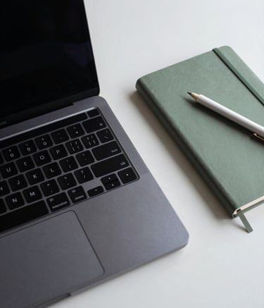 A top-down view of a minimalist workstation featuring a dark navy laptop, a sage green notebook, and professional pearl white stationary on a clean desk.