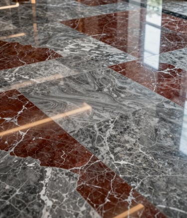High-angle photography of a luxury hotel lobby floor featuring large, polished stone grey and deep crimson marble tiles with intricate natural patterns. The surface is highly reflective, showing high-end lighting and the robust, expert craftsmanship of the stone installation.