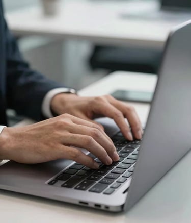 A close-up photograph of a professional's hands working on a sleek black laptop in a sophisticated North American marketing firm. The setting is bright and clean with white surfaces and grey accents. High-quality lighting emphasizes a focused and innovative digital work environment.