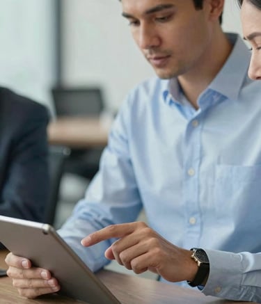 A close-up photograph of a professional consultant in a soft sky blue shirt pointing at a tablet screen during a meeting. The background shows a blurred, modern office environment with a professional atmosphere.