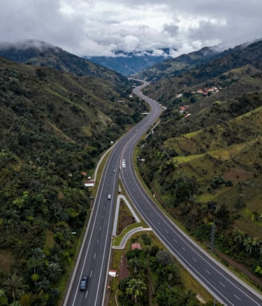 An aerial wide-angle shot of a newly constructed highway winding through a lush mountain landscape. Modern Latin American / Spanish infrastructure. Crisp, high-resolution photography with mist-colored clouds and dark navy asphalt tones.