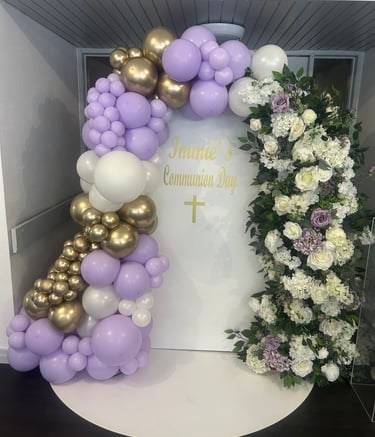 Purple, white, and gold balloon arch with floral backdrop for a First Holy Communion celebration.