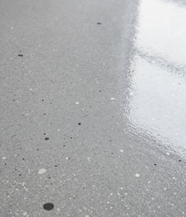 A close-up, high-angle shot of a cured epoxy floor with a decorative flake finish in a palette of grey, charcoal, and white. The surface has a high-gloss, crystal-clear topcoat that reflects soft overhead lighting, showcasing a non-slip, professional-grade texture that embodies the #F7F9FB and #2B2F33 brand colors.